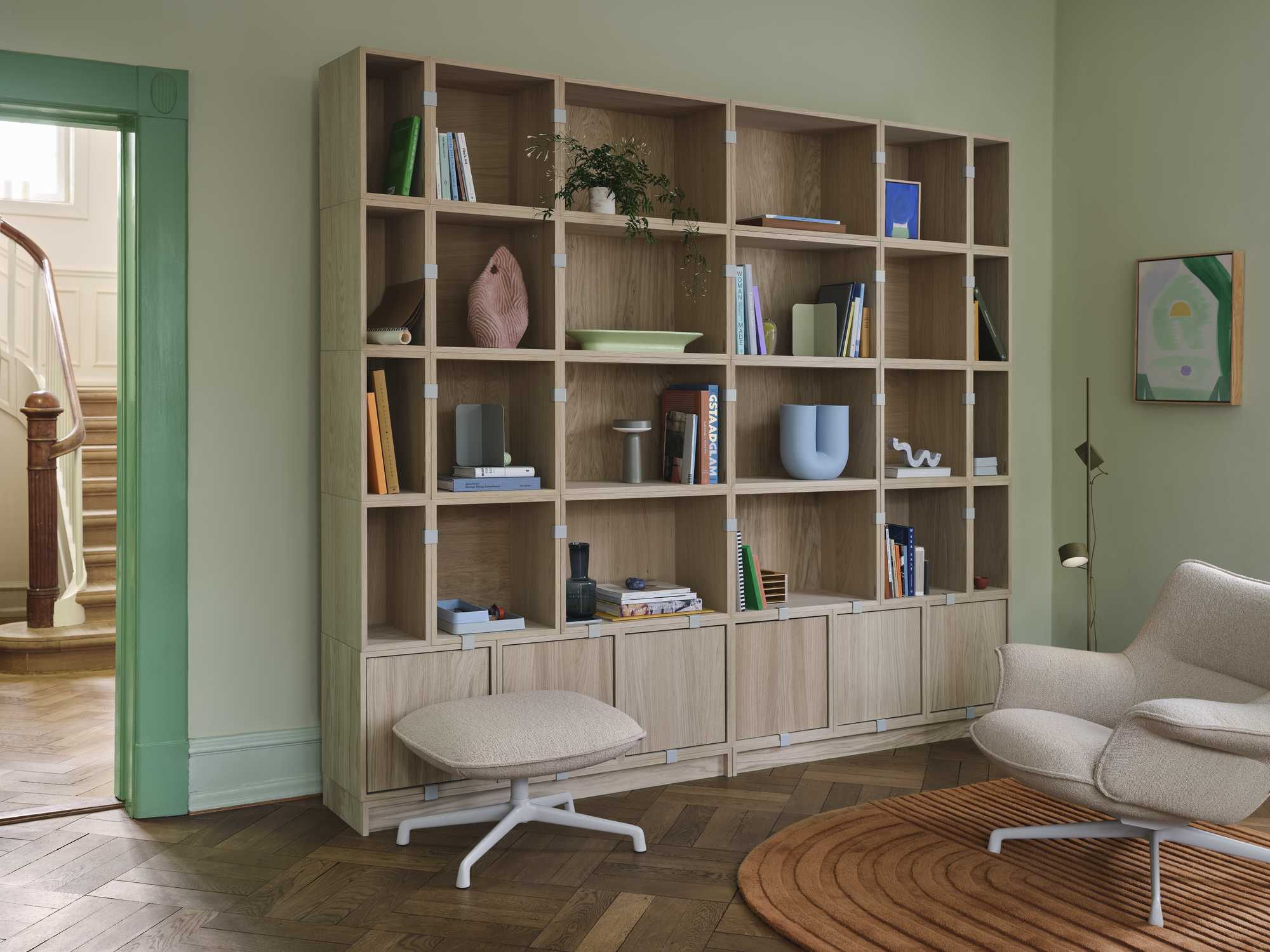 Stacked Storage System Bookcase Configuration 1 in Oak - Doze Lounge Chair Low Back & Ottoman Swivel Base in Hearth 7/Grey - Relevo Rug in Burnt Orange - Post Floor Lamp in Brown Green - Kink Vase in Light Blue - Ease Portable Lamp in Grey - Mere Bowl in Light Green - Raise Carafe in Dark Blue - Compile Bookend in Grey - Arrange Desktop Series in Light Blue